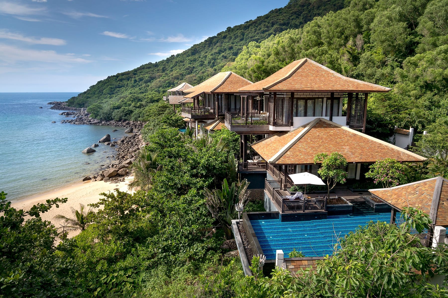Thai-roofed villas on a green hillside overlooking a turquoise bay